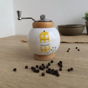 A small, round vintage-style pepper mill with a white porcelain body and a light wooden top. The mill features a subtle yellow geometric graphic on its side and has an exposed metal grinding mechanism at the top. The image is a close-up, showcasing the mill's design and internal structure, resting on a rustic farmhouse wooden table.
