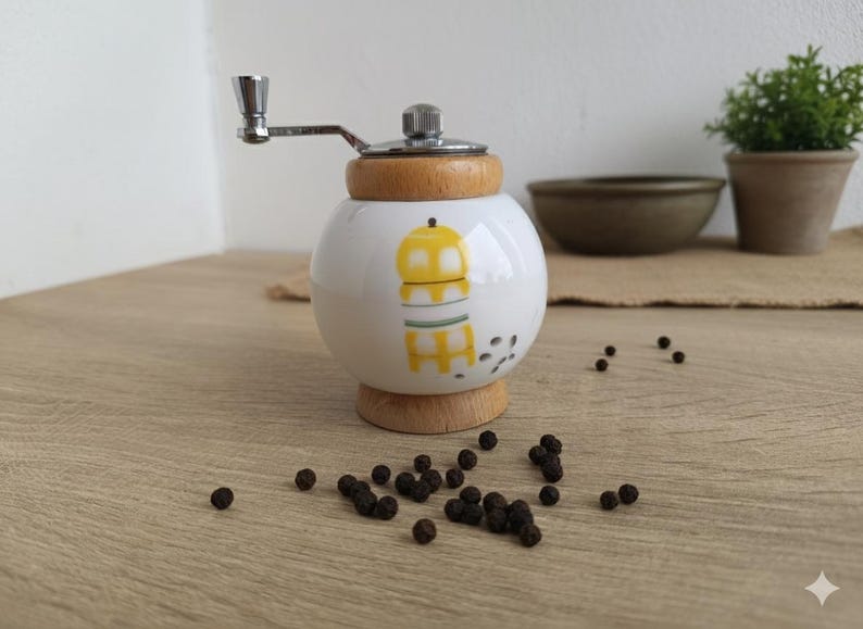 A small, round vintage-style pepper mill with a white porcelain body and a light wooden top. The mill features a subtle yellow geometric graphic on its side and has an exposed metal grinding mechanism at the top. The image is a close-up, showcasing the mill's design and internal structure, resting on a rustic farmhouse wooden table.