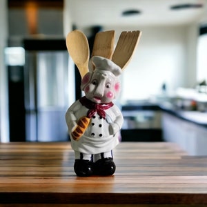Może przedstawiać: Biała ceramiczna figurka szefa kuchni trzymająca bochenek chleba, w czerwono-białym kapeluszu szefa kuchni i czerwonym szaliku. Figurka trzyma trzy drewniane narzędzia kuchenne: łyżkę, szpatułkę i drewnianą łyżkę.