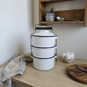 Op de afbeelding: Een witte geëmailleerde voedseldrager met drie niveaus, een zwarte handgreep en een blauwe rand. De drager staat op een houten oppervlak naast een patroon doek, een houten snijplank en een eierdoos.