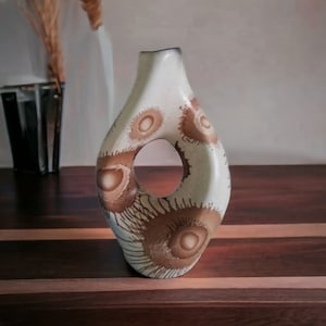 Puede incluir: Un jarrón de cerámica blanca con un diseño abstracto marrón y blanco. El jarrón tiene una forma única con una gran abertura en el centro.