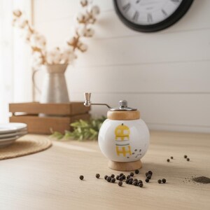 A small, round vintage-style pepper mill with a white porcelain body and a light wooden top. The mill features a subtle yellow geometric graphic on its side and has an exposed metal grinding mechanism at the top. The image is a close-up, showcasing the mill's design and internal structure, resting on a rustic farmhouse wooden table.