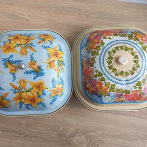 Op de afbeelding: Twee vintage metalen serveerschalen. Eén heeft een lichtblauwe achtergrond met een gele lelie bloemenontwerp. De andere heeft een beige achtergrond met een fruit- en bloemenontwerp. Beide hebben deksels en zijn vierkant met afgeronde hoeken.