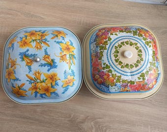Puede incluir: Dos fuentes de metal vintage. Una tiene un fondo azul claro con un diseño floral de lirios amarillos. La otra tiene un fondo beige con un diseño de frutas y flores. Ambas tienen tapas y son cuadradas con esquinas redondeadas.