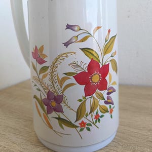 May include: White vintage thermos with a handle, featuring a floral design. The flowers are red, purple, and pink, with yellow and green leaves. The thermos is on a wooden surface.