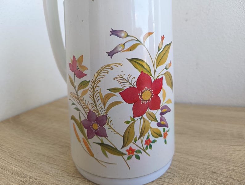 May include: White vintage thermos with a handle, featuring a floral design. The flowers are red, purple, and pink, with yellow and green leaves. The thermos is on a wooden surface.