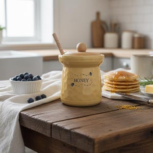 May include: A yellow ceramic honey pot with a wooden lid and spoon, with the word "HONEY" and bee illustrations. The pot is on a wooden table with blueberries, pancakes, and butter. A kitchen scene.