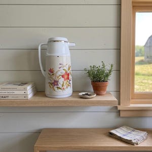 Vintage cream-colored thermos jug with a vibrant retro floral print featuring a red flower, purple blossoms, and wheat. The insulated carafe has a white plastic handle, lid, and pouring spout, accented by a shiny chrome metal trim. Inside, a reflective silver glass vacuum flask is visible. The white plastic screw-top lid has a small cream push button on top. The base is white with a large black plastic central screw underneath holding the flask secure
