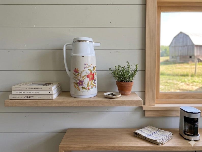 Vintage cream-colored thermos jug with a vibrant retro floral print featuring a red flower, purple blossoms, and wheat. The insulated carafe has a white plastic handle, lid, and pouring spout, accented by a shiny chrome metal trim. Inside, a reflective silver glass vacuum flask is visible. The white plastic screw-top lid has a small cream push button on top. The base is white with a large black plastic central screw underneath holding the flask secure