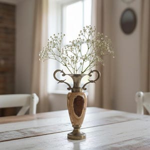 Op de afbeelding: Een decoratieve vaas met een lichtbruine en gouden basis, een lichtbruin en donkerbruin midden en zilveren handvatten. De vaas bevat een boeket kleine witte bloemen. De vaas staat op een witte houten tafel.