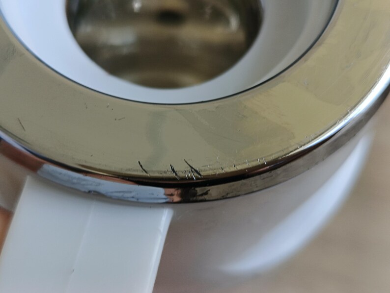 May include: Close-up of a white and silver tea kettle. The silver rim shows minor scratches. The white handle is visible on the bottom left.