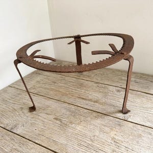 May include: A rusty, antique metal trivet with a circular top and three legs. The top has a serrated edge and three upward-pointing supports. The trivet is made of iron and has a weathered, aged appearance.