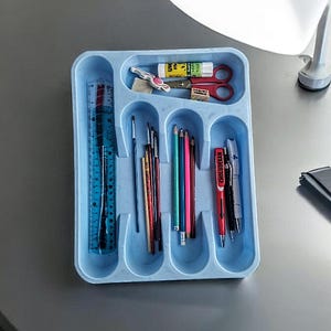 K&ouml;nnte beinhalten: Ein blaues Plastiktablett mit f&uuml;nf F&auml;chern enth&auml;lt verschiedene Schulmaterialien, darunter ein Lineal, Bleistifte, Stifte und Pinsel.