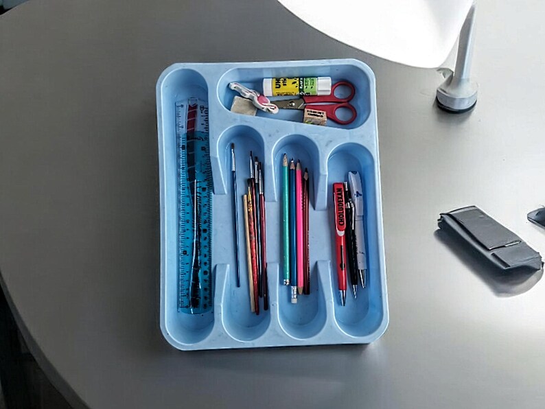 K&ouml;nnte beinhalten: Ein blaues Plastiktablett mit f&uuml;nf F&auml;chern enth&auml;lt verschiedene Schulmaterialien, darunter ein Lineal, Bleistifte, Stifte und Pinsel.