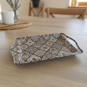 Könnte beinhalten: Rechteckiges Stofftablett mit detailliertem Muster aus Rauten und floralen Motiven in Braun-, Blau- und Cremetönen. Das Tablett liegt auf einem hellen Holztisch, was auf einen dekorativen oder funktionalen Zweck hindeutet.