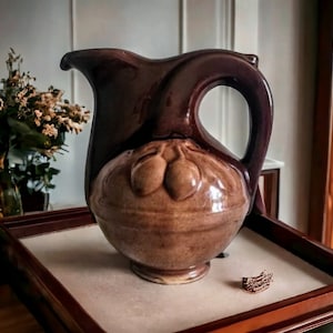 May include: A brown ceramic pitcher with a rounded body and a curved handle. The pitcher has a decorative design of three acorns on the front.