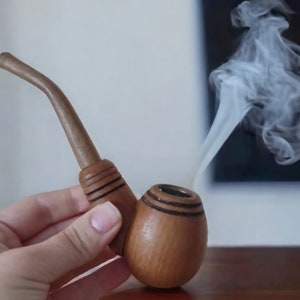 Può includere: Una pipa di legno con finitura marrone e nera, tenuta in una mano con fumo che sale dalla ciotola.
