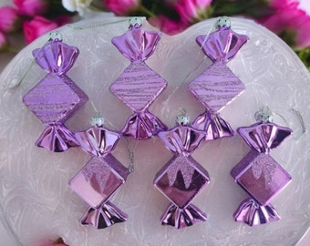 May include: Six purple candy-shaped Christmas ornaments with silver string hangers. The ornaments have a diamond shape in the centre with glitter accents and a shiny, wrapped candy design on the ends. The ornaments are displayed on a white, decorative plate.