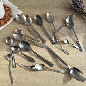Puede incluir: Una variedad de cucharas de acero inoxidable de varios tamaños, desde cucharaditas hasta cucharas de servir grandes, dispuestas sobre una superficie de madera clara. También hay una taza blanca y un ramo de lavanda seca.