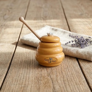May include: A light yellow ceramic honey pot with a wooden dipper, featuring a bee graphic and the words "honey miel". It rests on a wooden surface, alongside a linen cloth and lavender sprigs.