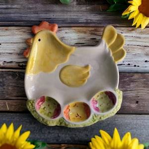 Puede incluir: Un plato de cerámica con forma de pollo con tres hendiduras en forma de huevo. El pollo es blanco con detalles amarillos y las hendiduras están decoradas con motivos florales verdes, rosas y amarillos.
