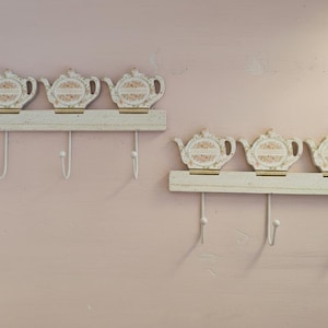 Op de afbeelding: Twee sets decoratieve wandhaken. Elke set heeft een witte houten basis met drie metalen haken en drie theepotvormige ornamenten. De theepotten hebben een bloemenontwerp en zijn geschilderd in zachte pastelkleuren.