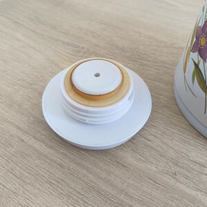 May include: White plastic lid with a central hole and a tan-colored seal, resting on a light wooden surface. The lid is from a white ceramic container with floral designs in purple, yellow, and red.