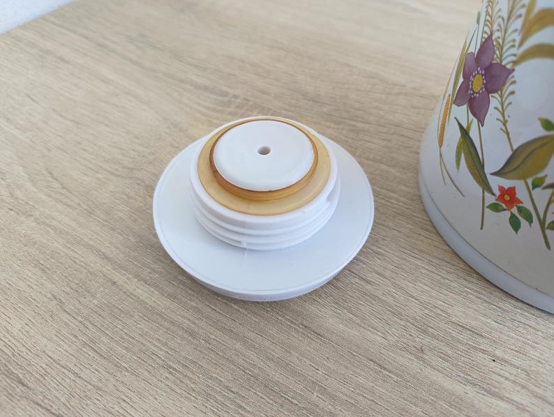 May include: White plastic lid with a central hole and a tan-colored seal, resting on a light wooden surface. The lid is from a white ceramic container with floral designs in purple, yellow, and red.