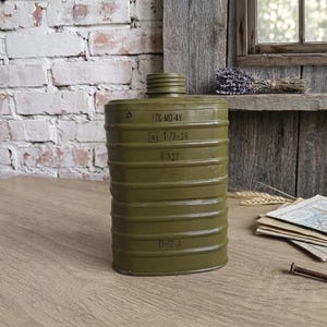 May include: An olive green metal gas mask filter canister with a threaded top opening. The cylindrical body features horizontal ridges and printed markings including FCC-MD-4Y, 1-72-36, B-322, and E1-12-A. The surface has a smooth, matte finish.