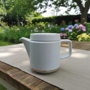 May include: A white ceramic teapot with a matching lid, placed on a wooden table with a beige linen runner. The teapot has a gold band at the base and a square handle. The background shows a garden with flowers.