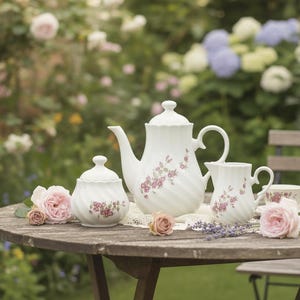Könnte beinhalten: Ein weißes Porzellan-Teeservice mit Teekanne, Zuckerdose, Sahnekännchen und Teetasse, verziert mit rosa Blumenmustern. Das Set steht auf einem Holztisch mit Rosen und Lavendelzweigen, vor einem verschwommenen Gartenhintergrund.