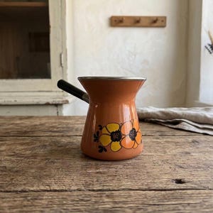 Può includere: Una caffettiera in smalto arancione con manico nero e un motivo floreale giallo e arancione. La pentola ha un bordo argentato e poggia su una superficie di legno rustico. Un oggetto vintage per la cucina.