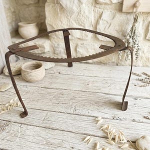 May include: A rustic, brown metal trivet with a circular, serrated top and three curved legs. The trivet is designed to hold hot cookware and protect surfaces. The design has an aged, antique appearance, suggesting it is vintage or handcrafted.