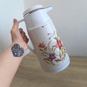 May include: White vintage thermos with a floral design featuring red, purple, and yellow flowers. The thermos has a white handle and a silver rim. The floral pattern is on a cream-colored background.