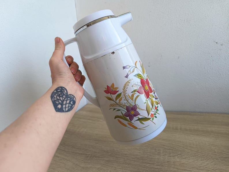 May include: White vintage thermos with a floral design featuring red, purple, and yellow flowers. The thermos has a white handle and a silver rim. The floral pattern is on a cream-colored background.