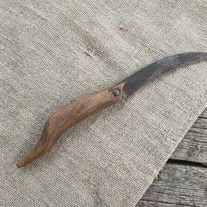 May include: A vintage curved blade knife with a wooden handle. The blade is dark and shows signs of wear, while the handle is a light brown color. The knife is resting on a textured, tan fabric, with a wooden surface in the background.