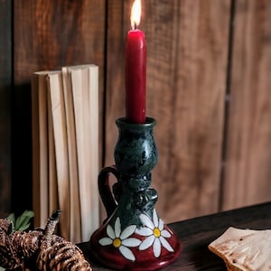 May include: A ceramic candle holder with a green and blue glaze and white daisy accents. The holder has a red base and a red candle is lit inside.