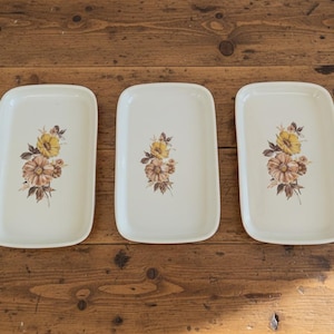 Op de afbeelding: Drie rechthoekige serveerschalen met afgeronde hoeken. Elke schaal is gebroken wit en heeft een bloemenontwerp met gele, perzik- en bruine bloemen. De schalen zijn gerangschikt op een rustiek houten oppervlak.