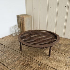 Puede incluir: Un salvamanteles redondo de metal oxidado con una parte superior de listones y tres patas cortas. El salvamanteles está diseñado para proteger las superficies del calor. Se coloca sobre una superficie de madera envejecida, con libros y botellas de vidrio al fondo.