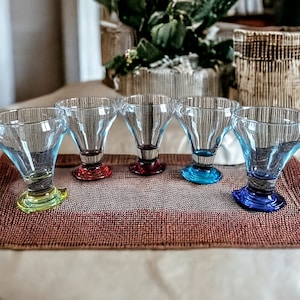 May include: Six clear glass dessert cups with colorful bases. The bases are red, blue, green, and yellow. The cups are arranged in a row on a brown placemat.