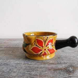 May include: A ceramic sauce boat with a mustard yellow base colour, featuring a hand-painted red floral design with black outlines. The vessel includes a glossy black handle extending from the side and a small pouring spout on the rim.