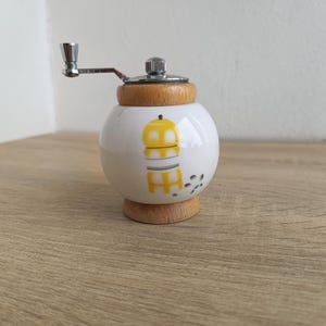 A small, round vintage-style pepper mill with a white porcelain body and a light wooden top. The mill features a subtle yellow geometric graphic on its side and has an exposed metal grinding mechanism at the top. The image is a close-up, showcasing the mill's design and internal structure, resting on a rustic farmhouse wooden table.