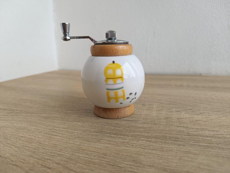 A small, round vintage-style pepper mill with a white porcelain body and a light wooden top. The mill features a subtle yellow geometric graphic on its side and has an exposed metal grinding mechanism at the top. The image is a close-up, showcasing the mill's design and internal structure, resting on a rustic farmhouse wooden table.