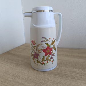 May include: A vintage cream-colored thermos with a white top and handle. The thermos features a floral design with red, purple, and green flowers. The spout is silver-colored.