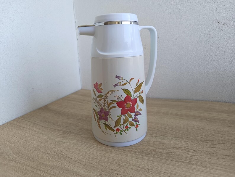 May include: A vintage cream-colored thermos with a white top and handle. The thermos features a floral design with red, purple, and green flowers. The spout is silver-colored.