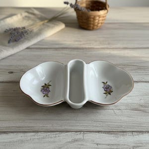 Op de afbeelding: Wit verdeeld serveerschaaltje met een bloemmotief. De schaal heeft twee afgeronde secties en een centrale, smalle sectie. Elke afgeronde sectie heeft een paarse bloem met groene bladeren. De schaal heeft een bruine rand.