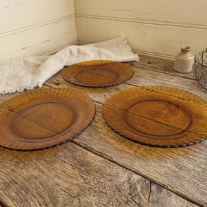 May include: Three amber-colored glass plates with a textured, ridged rim and a wood-grain center are displayed on a rustic wooden surface. A linen napkin, a small ceramic bottle, and a wire basket are also present.