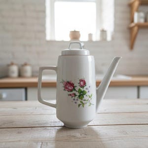 May include: White ceramic coffee pot with a floral rose design. The pot has a rectangular handle, a long spout, and a lid. The floral design features pink roses and green leaves. The pot is on a light-coloured wooden table.