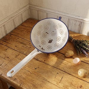 May include: A white enamel colander with a blue rim and handle rests on a wooden surface. The colander has numerous small holes. A small bundle of dried herbs, two potatoes, and an eggshell are also on the table.