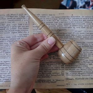 Può includere: Primo piano di una pipa di legno con un design a strisce marroni e bianche. La pipa è tenuta da una mano sopra un libro con testo.